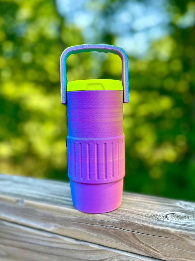 Puede incluir: Una botella de agua vibrante con asa y dise&ntilde;o degradado. La botella presenta una transici&oacute;n de morado a azul, con una tapa verde ne&oacute;n y un asa que se desvanece de azul a verde. Tiene una secci&oacute;n acanalada para el agarre.