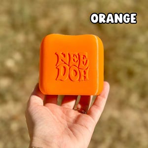 May include: A bright orange, square-shaped Nee Doh stress toy with rounded corners. The word "NEE DOH" is embossed in a stylized, bold font on the front surface of the object. The toy has a smooth, solid-colored finish.