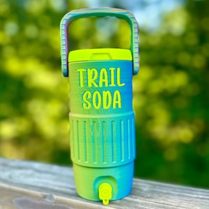 Puede incluir: Un enfriador de agua colorido con asa, con un degradado de verde y azul. Las palabras "TRAIL SODA" est&aacute;n impresas en la parte delantera. Tiene un grifo y una tapa amarillos, perfectos para actividades al aire libre.