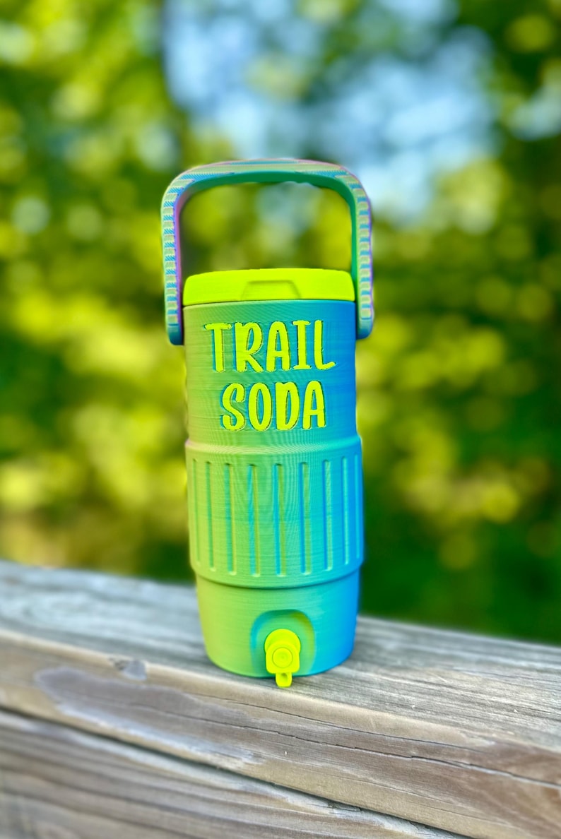 Puede incluir: Un enfriador de agua colorido con asa, con un degradado de verde y azul. Las palabras "TRAIL SODA" est&aacute;n impresas en la parte delantera. Tiene un grifo y una tapa amarillos, perfectos para actividades al aire libre.