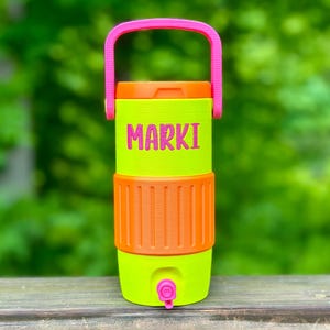 May include: A neon green and orange water cooler with a pink handle. The word "MARKI" is printed in pink on the front. The cooler has a spigot at the bottom and is designed for outdoor use.