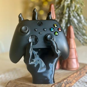 Può includere: Controller Xbox nero tenuto da un supporto a forma di mano nera, appoggiato su una base di legno. Il controller ha pulsanti colorati. Alberi di Natale decorativi color rame e verdi sullo sfondo.