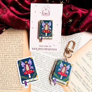 Op de afbeelding: Een harde emaille pin, sticker en sleutelhanger met een feeënontwerp op een boek. De fee heeft paarse vleugels en zit op een rode paddenstoel. Het boek is teal met gouden details.