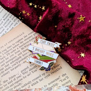 May include: A white enamel pin with a stack of colorful books and the text "Plot Twist I Bought More Books". The pin is on a burgundy velvet background with gold stars.