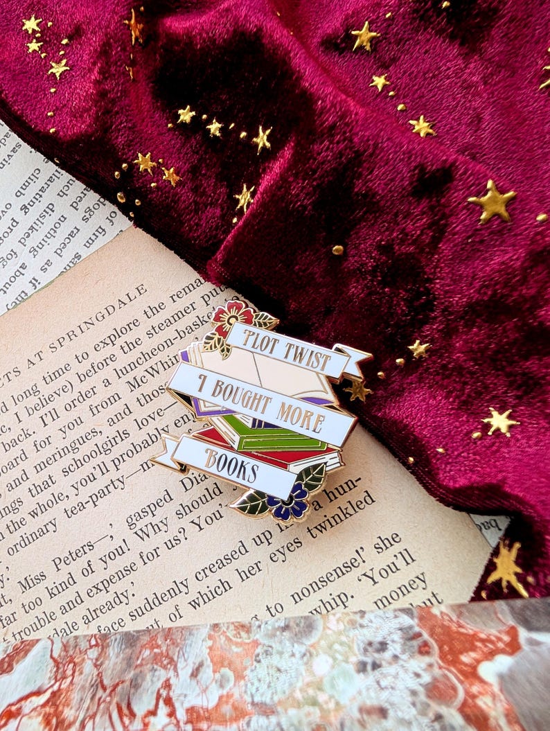 May include: A white enamel pin with a stack of colorful books and the text "Plot Twist I Bought More Books". The pin is on a burgundy velvet background with gold stars.
