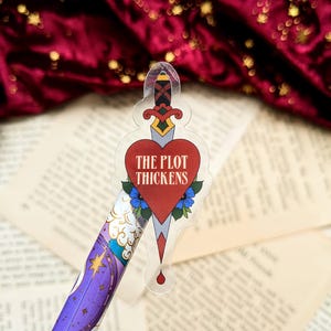 May include: A clear sticker with a red heart pierced by a dagger. The heart has the text "THE PLOT THICKENS" inside.