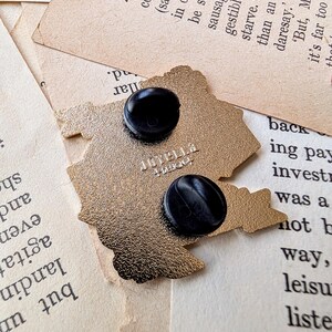 May include: Gold-colored metal pin featuring a cartoon image of a house with a chimney and a window. The pin has a black backing with a safety pin closure. The pin is on a white surface.