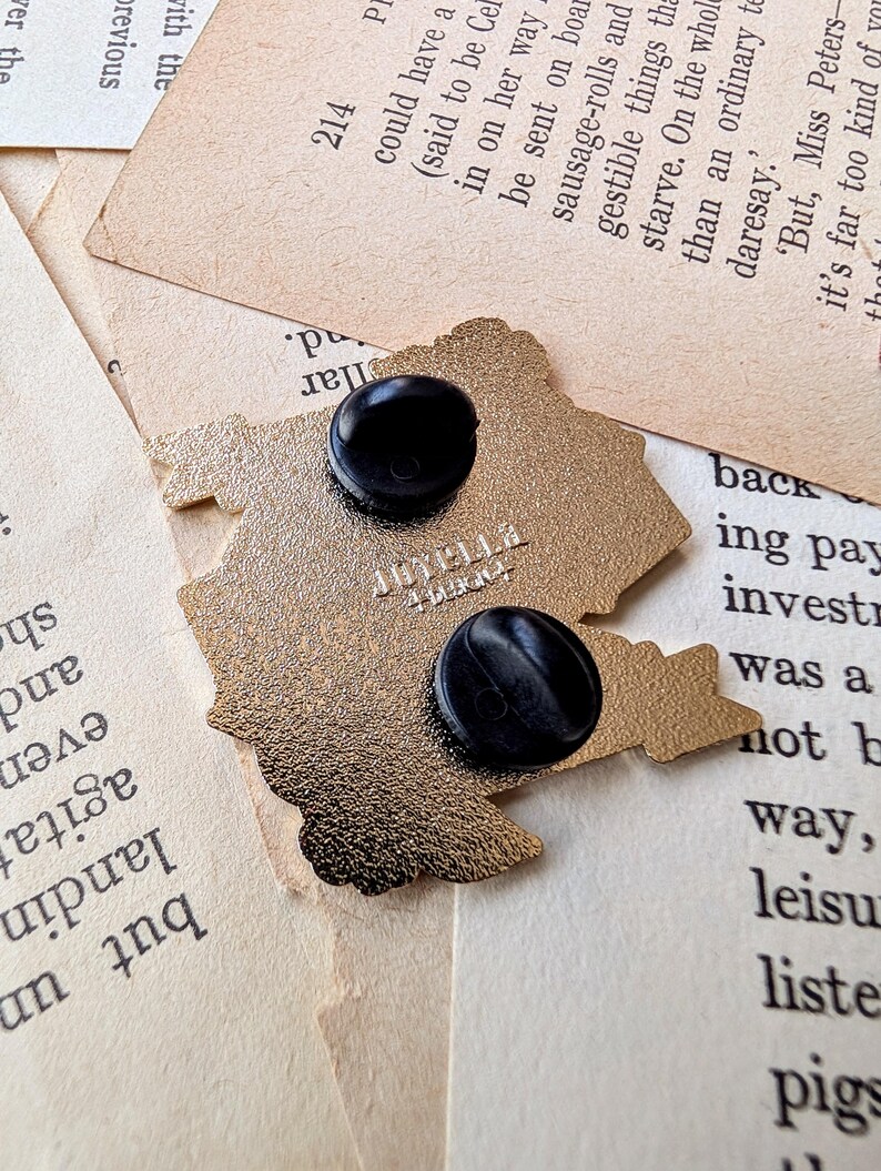 May include: Gold-colored metal pin featuring a cartoon image of a house with a chimney and a window. The pin has a black backing with a safety pin closure. The pin is on a white surface.