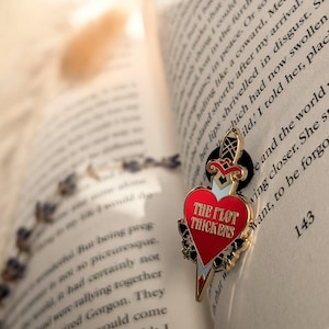 May include: A red heart-shaped enamel pin with a gold dagger design and the text "THE PLOT THICKENS." The pin is set against the pages of an open book, with a blurred background of dried flowers.