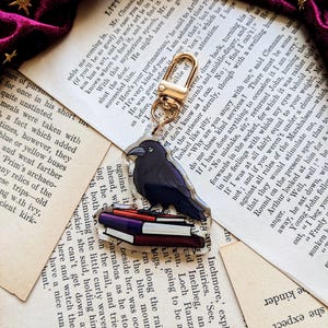 May include: A black raven charm with a stack of colourful books. The charm is attached to a gold key ring.