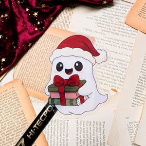 Pode incluir: Adesivo de um fantasma branco usando um chapéu e laço vermelhos de Papai Noel, segurando livros. O fantasma tem olhos grandes e pretos e um sorriso. O fundo inclui páginas de livros antigos e um tecido de veludo vermelho.
