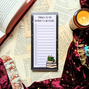Könnte beinhalten: Eine schwarz-weiße Checkliste mit dem Text "Things to do before I can read..." und einer Liste leerer Kreise. Ein grüner Drache sitzt auf einem Stapel Bücher. Der Hintergrund ist dunkelblau mit weißen Sternen.