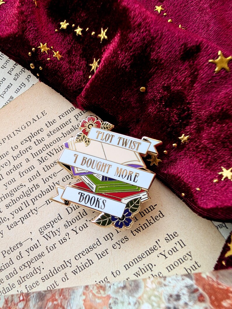 May include: Gold enamel pin with a stack of books and the text "I bought more books" and "Plot Twist".