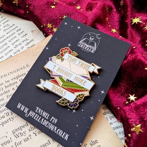 May include: A gold enamel pin with a stack of colorful books and a banner that reads "Plot Twist I Bought More Books". The pin is on a black background with a white skull and book logo that reads "Joyella Design".
