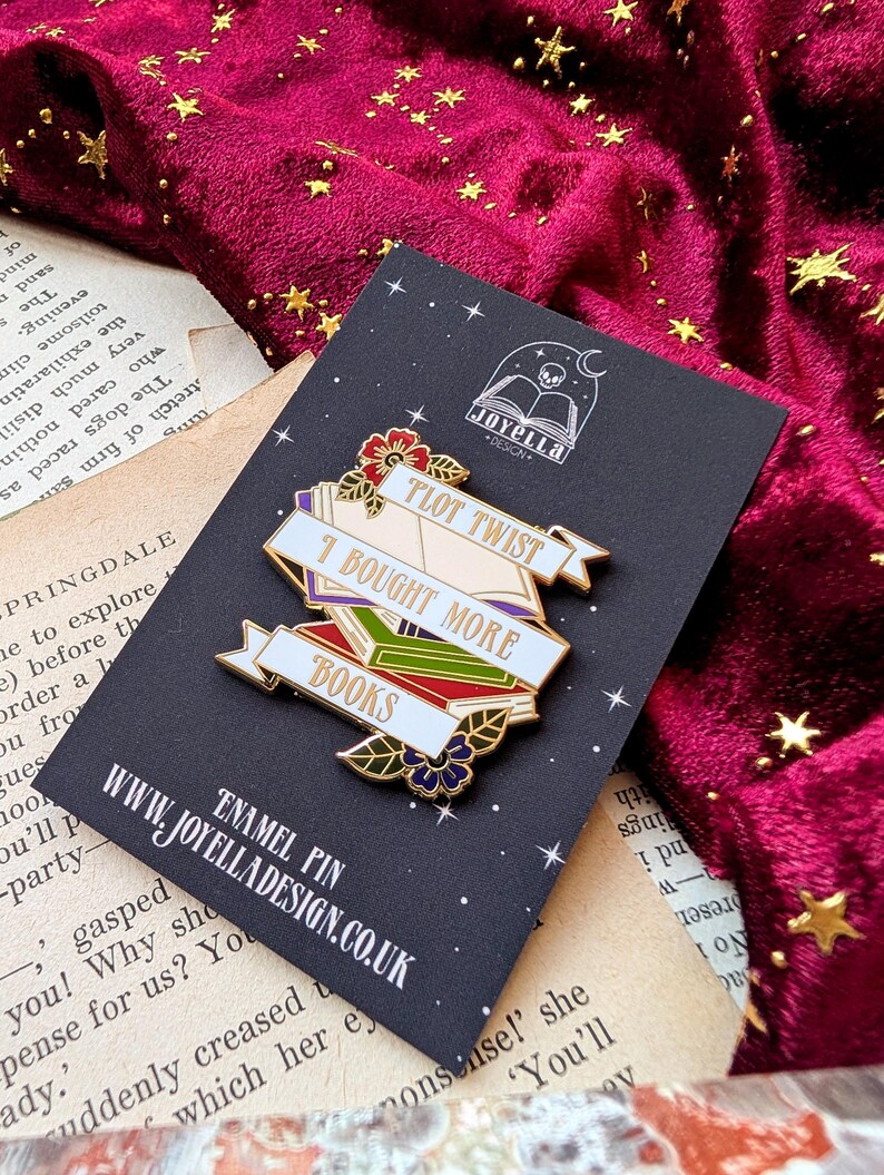 May include: A gold enamel pin with a stack of colorful books and a banner that reads "Plot Twist I Bought More Books". The pin is on a black background with a white skull and book logo that reads "Joyella Design".
