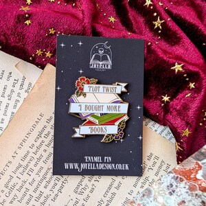 May include: A gold enamel pin with a white banner that reads "Plot Twist I Bought More Books". The banner is wrapped around a stack of colorful books with a flower and leaves on the bottom. The pin is on a black background with a skull and book logo that reads "Joyella Design".