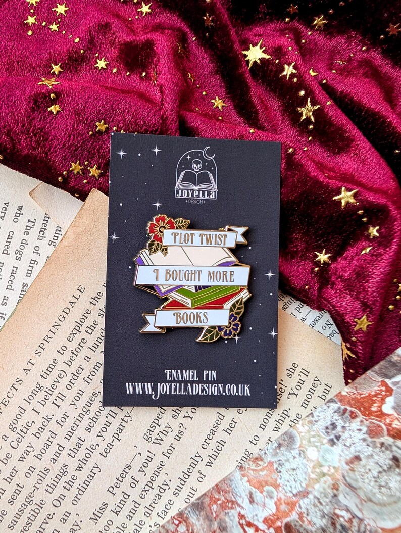 May include: A gold enamel pin with a white banner that reads "Plot Twist I Bought More Books". The banner is wrapped around a stack of colorful books with a flower and leaves on the bottom. The pin is on a black background with a skull and book logo that reads "Joyella Design".