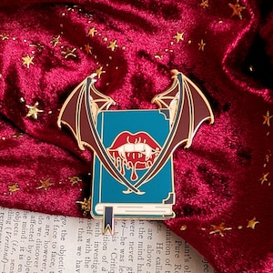 May include: A gold enamel pin depicting a blue book with a red mouth and fangs design, flanked by maroon bat wings. The pin is set against a red velvet background with gold stars.