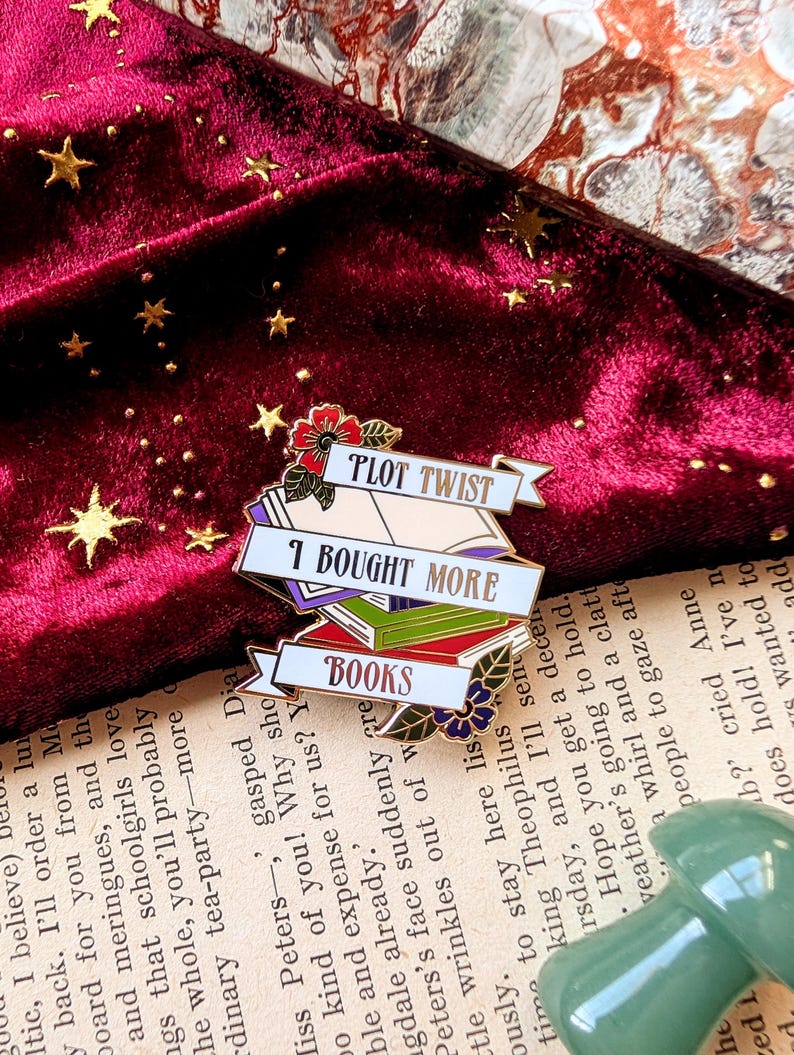 May include: Gold-tone enamel pin with a stack of colorful books, a banner that reads "Plot Twist: I Bought More Books", and a floral design.