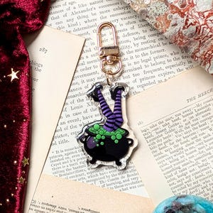 May include: A whimsical Halloween-themed keychain featuring a black cauldron with bubbling green potion and witch legs in striped purple and black stockings. The charm is attached to a gold-colored clasp.