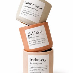 May include: Three stacked, cylindrical candles in neutral tones. Each candle has a white label with a definition: "entrepreneur," "girl boss," and "badassery." The candles are in shades of beige, peach, and light pink.