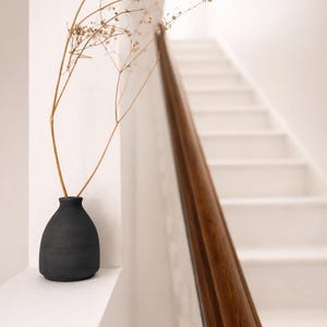 May include: A dark grey vase with dried flowers stands on a white ledge, with a staircase and wooden handrail in the background. The vase is a simple, rounded shape. The scene has a minimalist, neutral aesthetic.