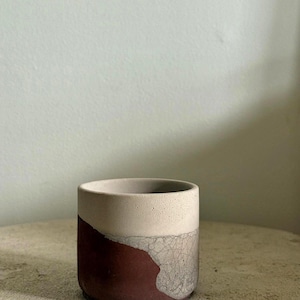May include: A two-tone concrete plant pot. The top section is light beige, and the lower section is a deep burgundy with a cracked, textured effect. Suitable for small plants or succulents. The pot is cylindrical in shape.