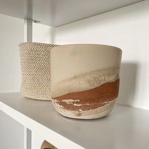 May include: A beige and brown decorative planter with a textured, layered design. The planter is cylindrical and sits on a white shelf next to a woven basket. The planter has a rustic, earthy aesthetic.