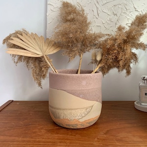 May include: A ceramic vase in shades of pink, beige, and brown, holding dried pampas grass and a fan-shaped palm leaf. The vase has a rounded shape and sits on a wooden surface. The vase is a decorative item for the home.