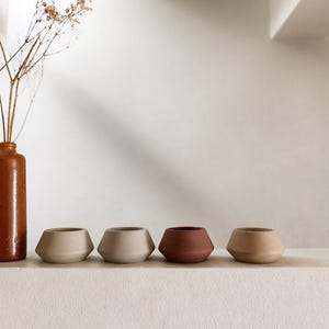 May include: Four small, geometric ceramic pots in beige and terracotta shades are arranged on a white shelf. A tall, brown ceramic vase with dried flowers is to the left. The pots have a modern, minimalist design.