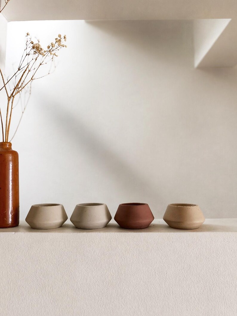 May include: Four small, geometric ceramic pots in beige and terracotta shades are arranged on a white shelf. A tall, brown ceramic vase with dried flowers is to the left. The pots have a modern, minimalist design.