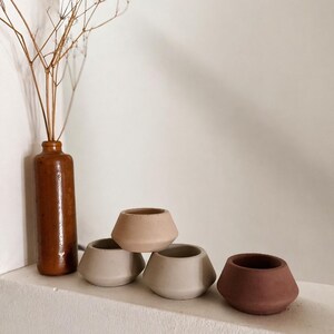 May include: A selection of small, geometric ceramic pots in beige and brown hues. A tall, brown bottle contains dried flowers. The pots are displayed on a white ledge, creating a minimalist, earthy aesthetic.