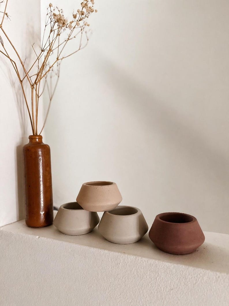 May include: A selection of small, geometric ceramic pots in beige and brown hues. A tall, brown bottle contains dried flowers. The pots are displayed on a white ledge, creating a minimalist, earthy aesthetic.