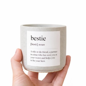 May include: A light grey ceramic candle holder with the word "bestie" printed on a white label. The label also includes a definition of "bestie". The candle holder is held in a hand.