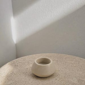 May include: A small, cream-coloured ceramic candle holder with a rounded base and a cylindrical opening. It rests on a textured, light-coloured surface. The background is a white wall with a shadow.