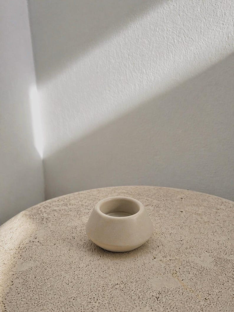 May include: A small, cream-coloured ceramic candle holder with a rounded base and a cylindrical opening. It rests on a textured, light-coloured surface. The background is a white wall with a shadow.
