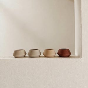 May include: Four small, geometric ceramic pots in a row. The pots are in shades of beige, tan, and reddish-brown. They have a modern, minimalist design and sit on a white ledge against a white wall.