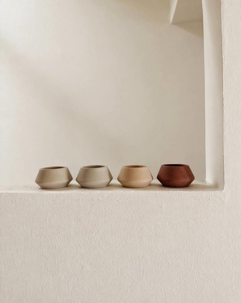 May include: Four small, geometric ceramic pots in a row. The pots are in shades of beige, tan, and reddish-brown. They have a modern, minimalist design and sit on a white ledge against a white wall.