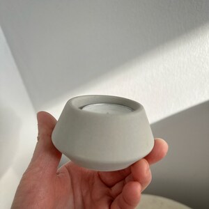 May include: A grey concrete candle holder with a geometric design. The holder has a wide base that tapers inwards to a smaller opening. A small, silver-coloured tea light candle is inside the holder. The holder is being held in a hand.