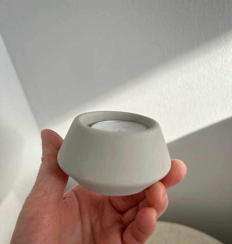 May include: A grey concrete candle holder with a geometric design. The holder has a wide base that tapers inwards to a smaller opening. A small, silver-coloured tea light candle is inside the holder. The holder is being held in a hand.