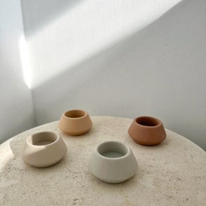 May include: Four small, ceramic candle holders in neutral tones: beige, light grey, peach, and terracotta. The holders have a rounded, asymmetrical shape and are arranged on a textured, light-coloured surface.