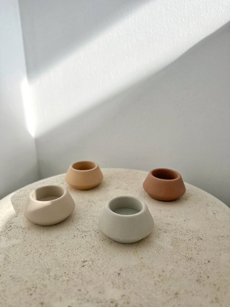 May include: Four small, ceramic candle holders in neutral tones: beige, light grey, peach, and terracotta. The holders have a rounded, asymmetrical shape and are arranged on a textured, light-coloured surface.
