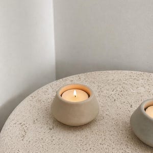 May include: A beige ceramic candle holder with a lit tea light candle. The holder has a rounded, organic shape and sits on a textured, light-coloured surface. The warm glow of the candle contrasts with the neutral tones.