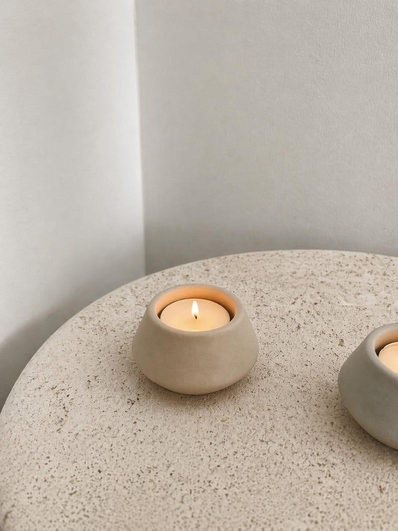 May include: A beige ceramic candle holder with a lit tea light candle. The holder has a rounded, organic shape and sits on a textured, light-coloured surface. The warm glow of the candle contrasts with the neutral tones.