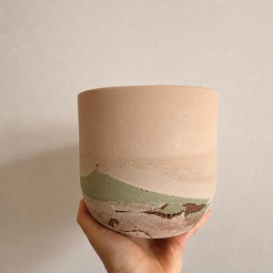 Peut inclure: Un pot de fleurs cylindrique avec une section supérieure lisse beige clair et une section inférieure texturée en couches, présentant des tons vert sauge et brun terreux. Le récipient a une base arrondie et une large ouverture supérieure.