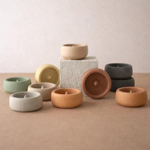 May include: A selection of small, round concrete candle holders in shades of green, grey, beige, and brown. Some are stacked, and one is inverted, showing the wick. The candle holders are displayed on a neutral-toned surface.