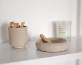 May include: A neutral-toned, minimalist aesthetic featuring a small, cylindrical container filled with wooden sticks and a round, shallow bowl holding a single stick. A candle is visible in the background. The items are arranged on a white shelf.