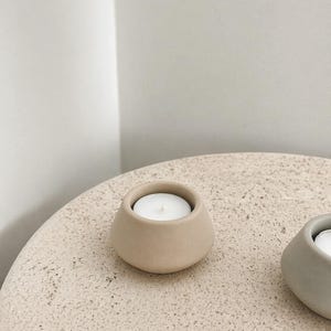 May include: A beige ceramic candle holder with a white candle inside. The holder has a rounded shape and sits on a textured, light-coloured surface. The candle holder is a simple, minimalist design.