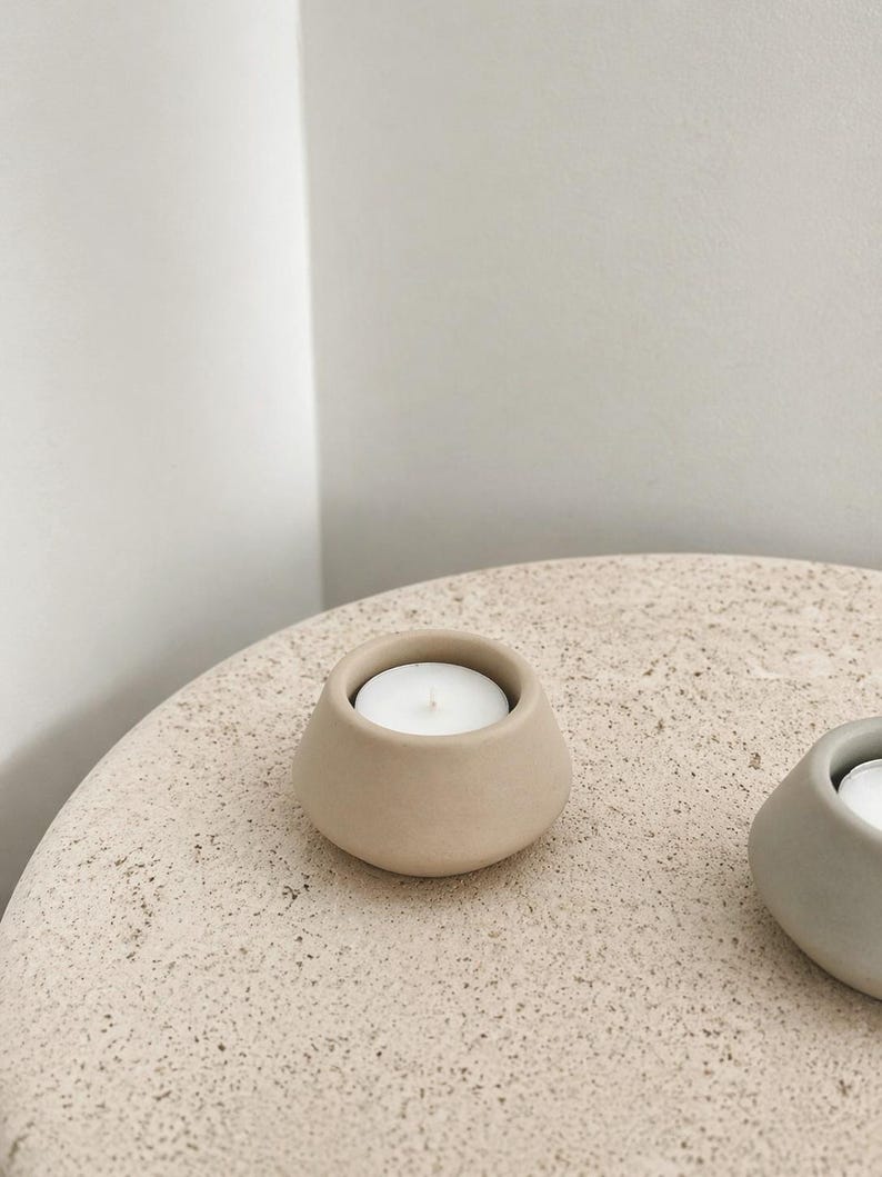 May include: A beige ceramic candle holder with a white candle inside. The holder has a rounded shape and sits on a textured, light-coloured surface. The candle holder is a simple, minimalist design.