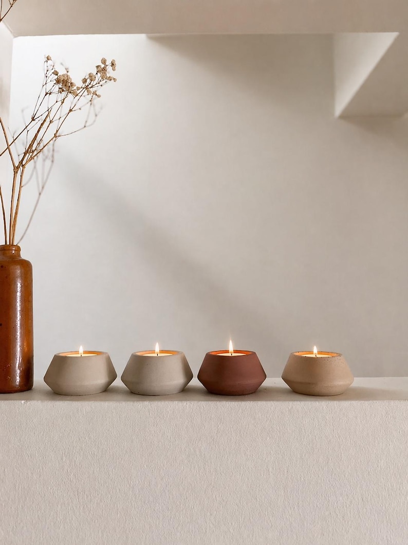 May include: Four lit candles in geometric, beige, and brown containers. The candles are arranged in a row on a white surface. A brown vase with dried flowers is on the left side of the image.